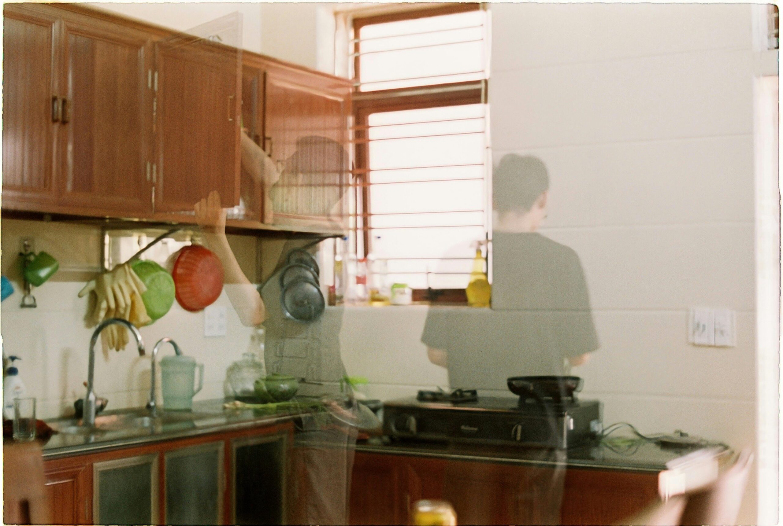 Fotografia de uma cozinha com efeito de múltipla exposição, apresentando tons quentes de madeira e luz natural vinda de uma janela com persiana ao fundo. A imagem sobrepõe dois momentos de uma pessoa em movimento: à esquerda, um vulto transparente alcança um armário suspenso sobre a pia; à direita, a mesma figura, também translúcida, aparece de costas próxima ao fogão. A composição sugere a passagem do tempo e a repetição de gestos no ambiente doméstico.  